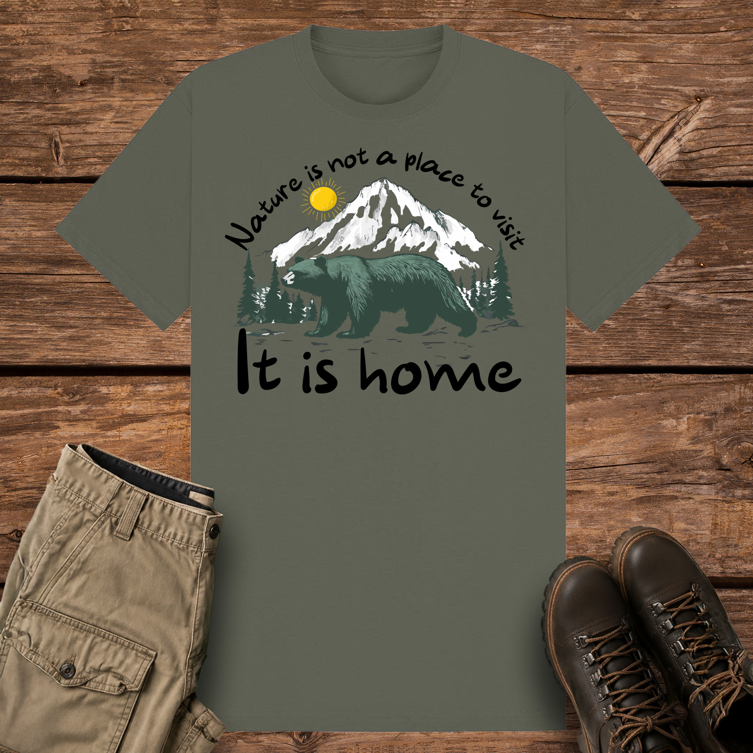 A khaki olive-colored graphic t-shirt featuring an illustration of a large dark green bear in front of snow-capped mountains and pine trees under a yellow sun. The text reads "Nature is not a place to visit" at the top and "It is home" at the bottom.