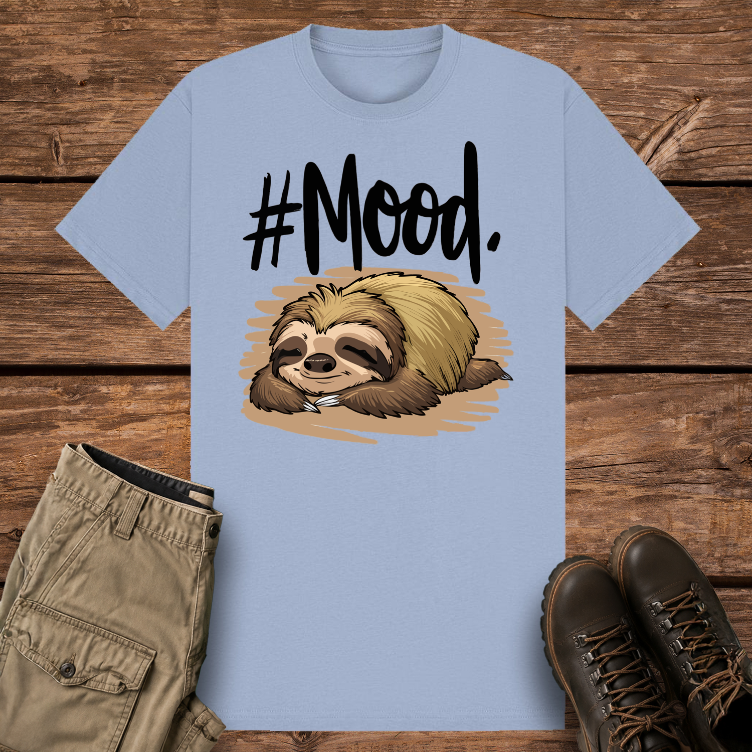 A pastel blue-colored graphic t-shirt featuring a detailed cartoon illustration of a brown and tan sloth sleeping flat on its stomach. Above the sloth, bold black brush-script text reads "#Mood."