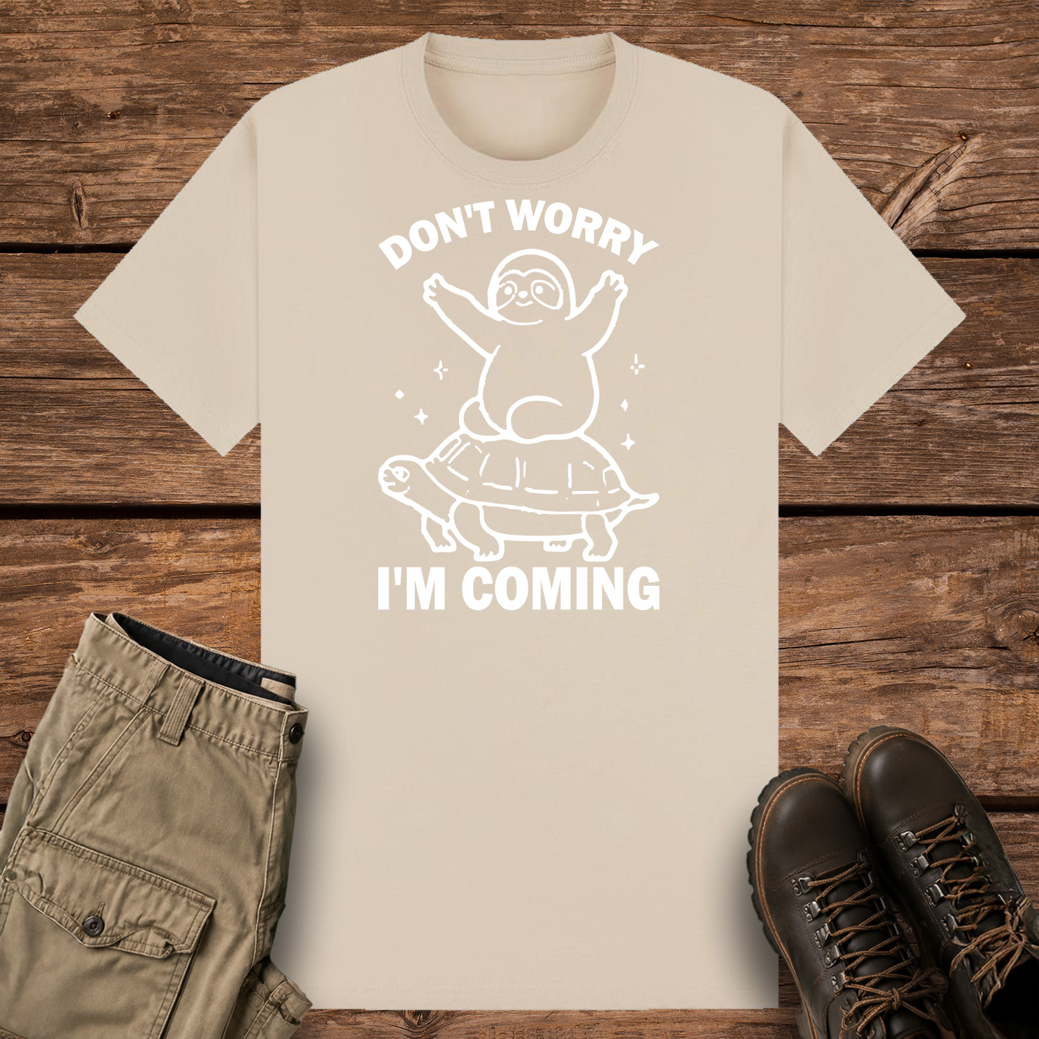 A sand graphic t-shirt featuring a minimalist white line art design of a cute cartoon sloth sitting on top of a sea turtle's shell. The text reads "DON'T WORRY" at the top and "I'M COMING" at the bottom.