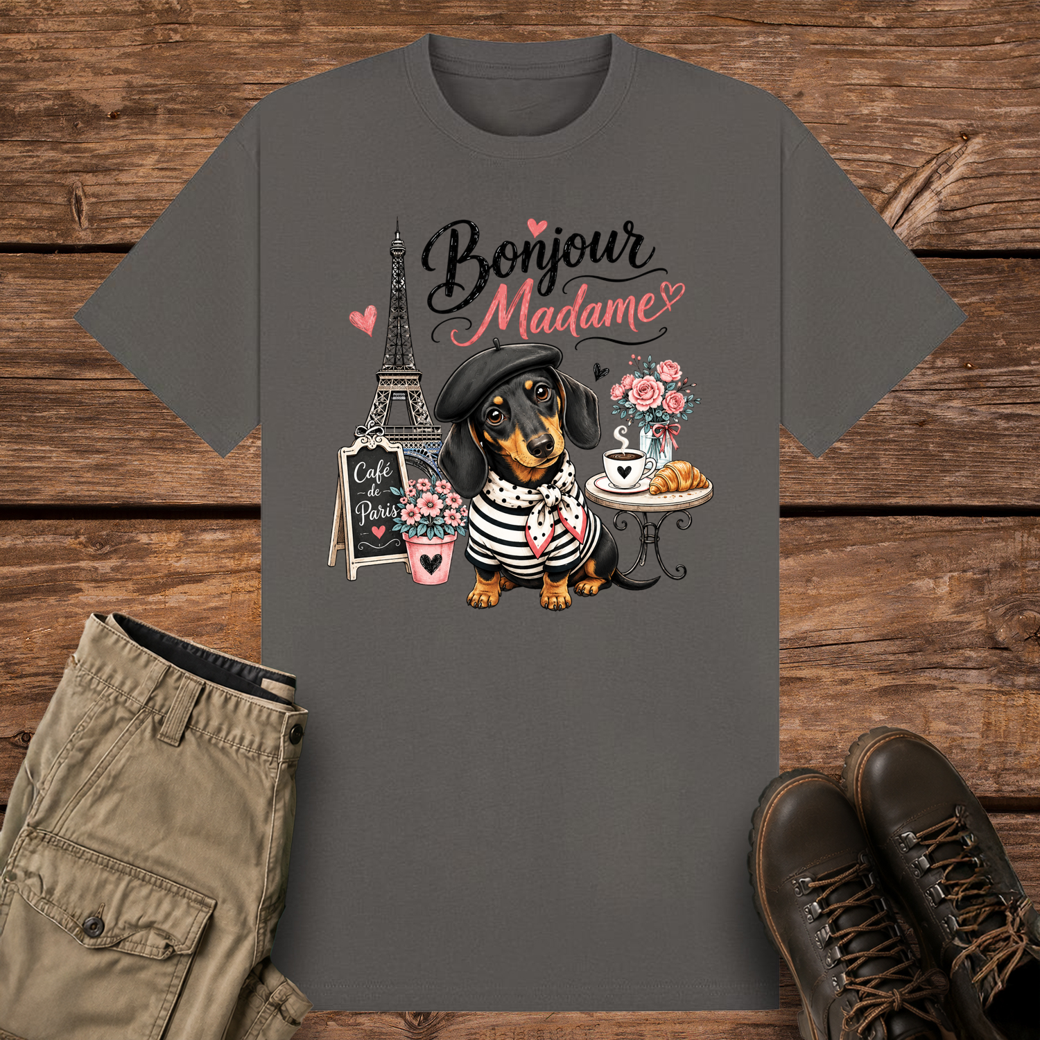 A charcoal-colored graphic t-shirt featuring a cute illustration of a black and tan dachshund dog wearing a beret and striped shirt in Paris. The design includes the Eiffel Tower, a café table with coffee, and the text "Bonjour Madame" with pink hearts.