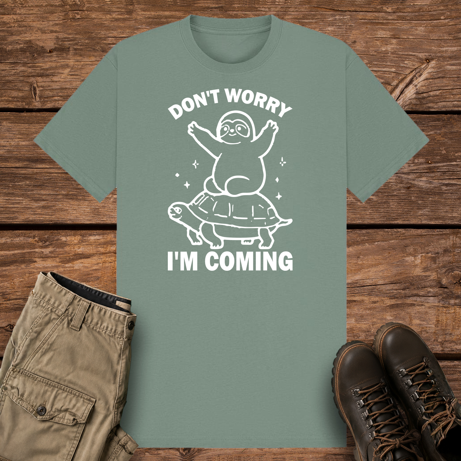 A sage graphic t-shirt featuring a minimalist white line art design of a cute cartoon sloth sitting on top of a sea turtle's shell. The text reads "DON'T WORRY" at the top and "I'M COMING" at the bottom.