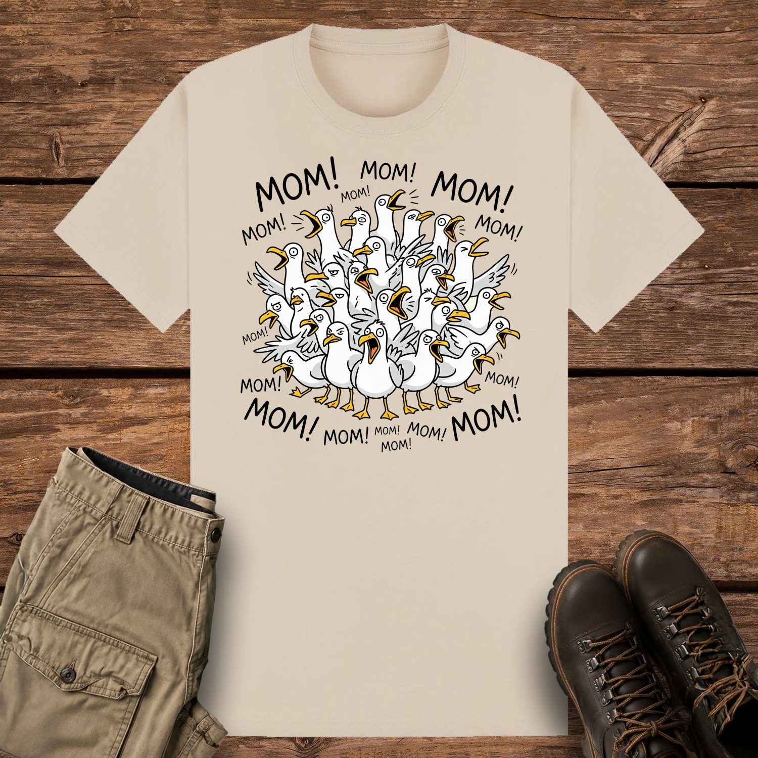 A sand-colored graphic t-shirt featuring a funny cartoon illustration of a crowded flock of squawking seagulls with their beaks wide open. The word "MOM!" is written repeatedly in a circle surrounding the frantic birds.