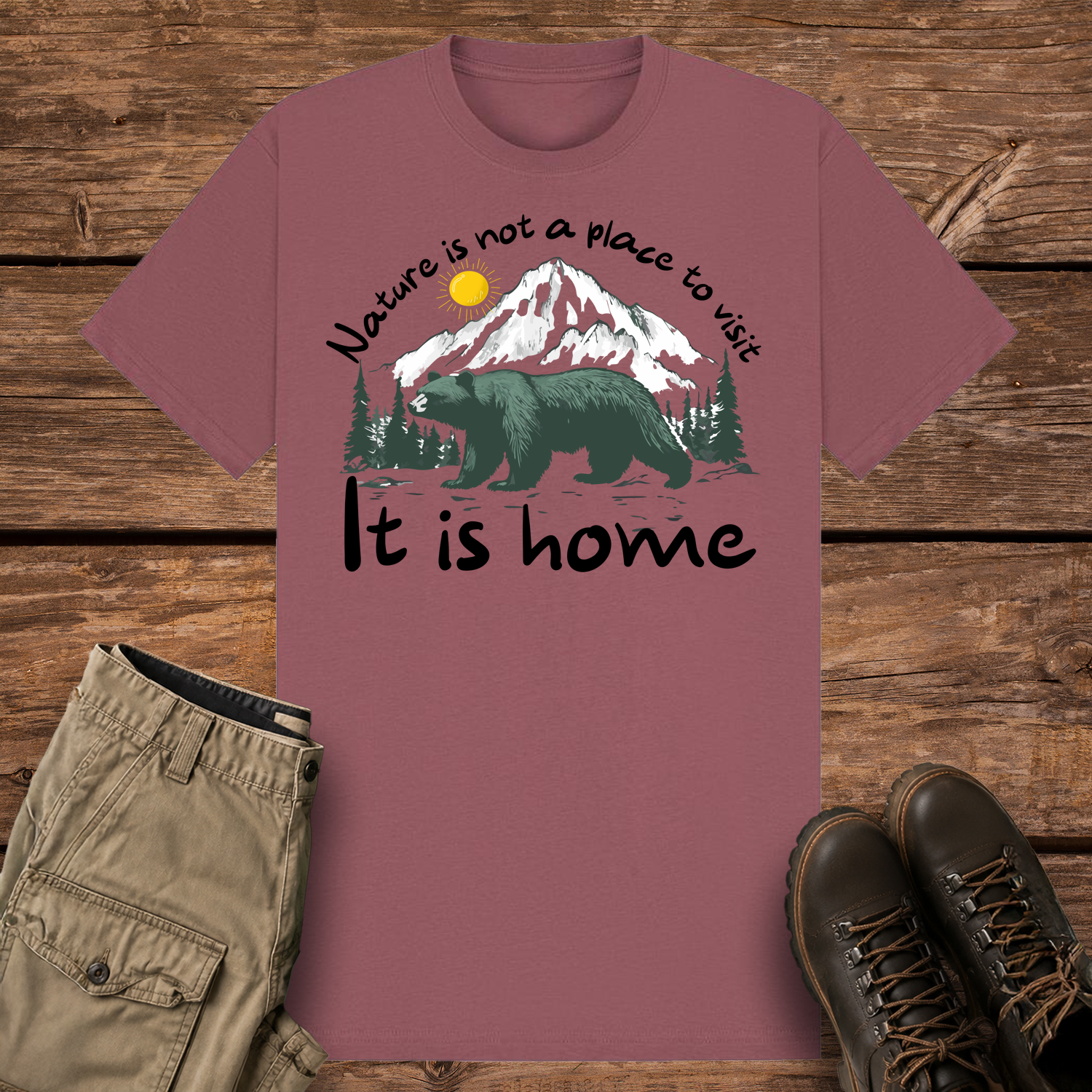 A mauve-colored graphic t-shirt featuring an illustration of a large dark green bear in front of snow-capped mountains and pine trees under a yellow sun. The text reads "Nature is not a place to visit" at the top and "It is home" at the bottom.