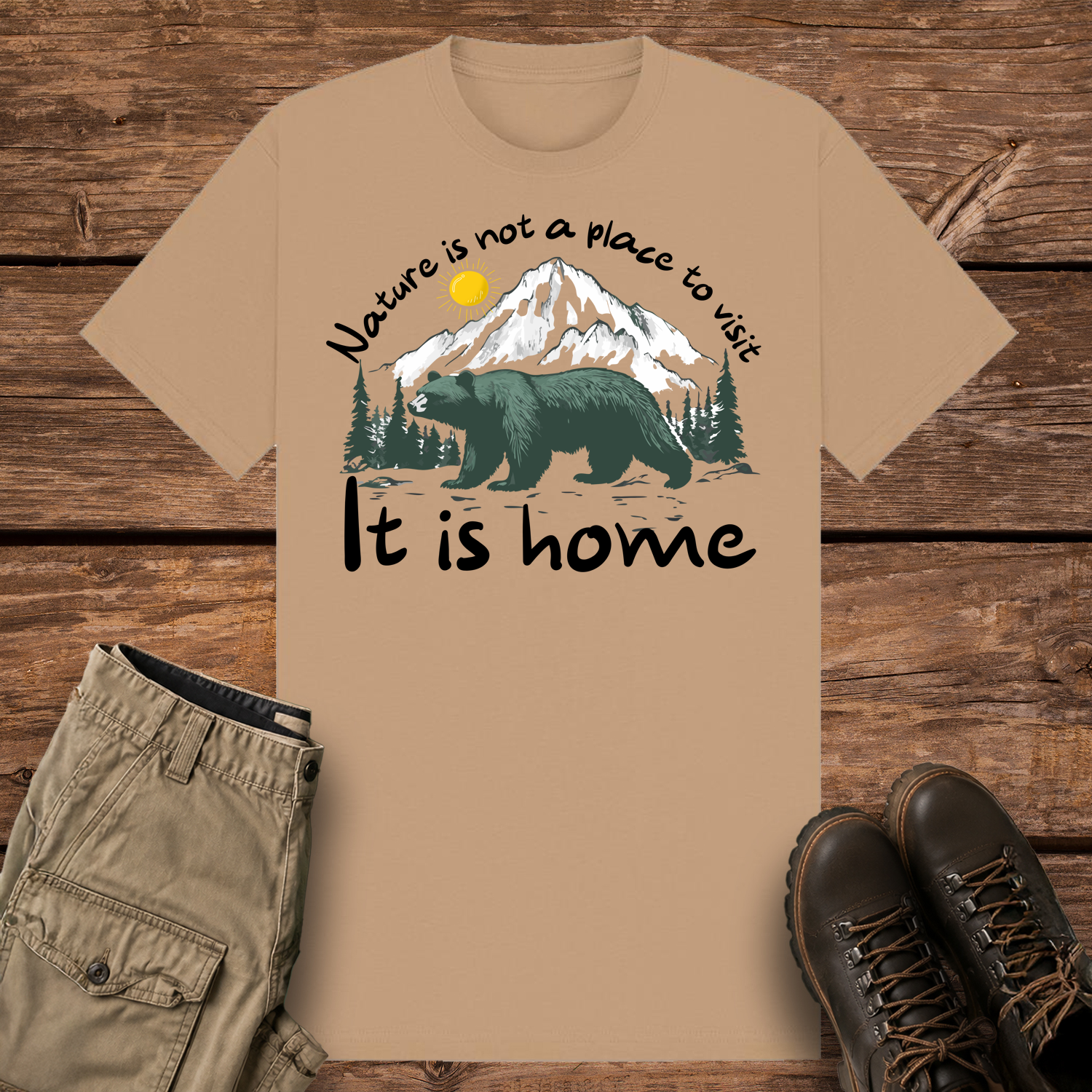 A camel-colored graphic t-shirt featuring an illustration of a large dark green bear in front of snow-capped mountains and pine trees under a yellow sun. The text reads "Nature is not a place to visit" at the top and "It is home" at the bottom.