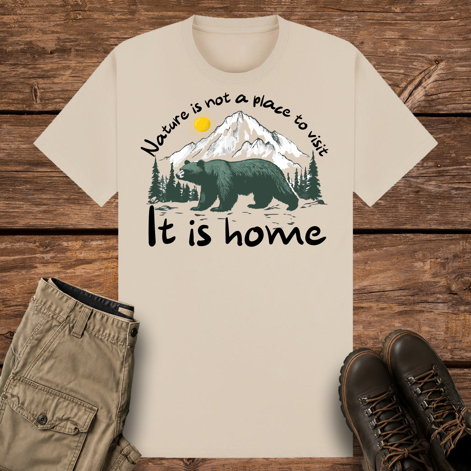 A sand-colored graphic t-shirt featuring an illustration of a large dark green bear in front of snow-capped mountains and pine trees under a yellow sun. The text reads "Nature is not a place to visit" at the top and "It is home" at the bottom.