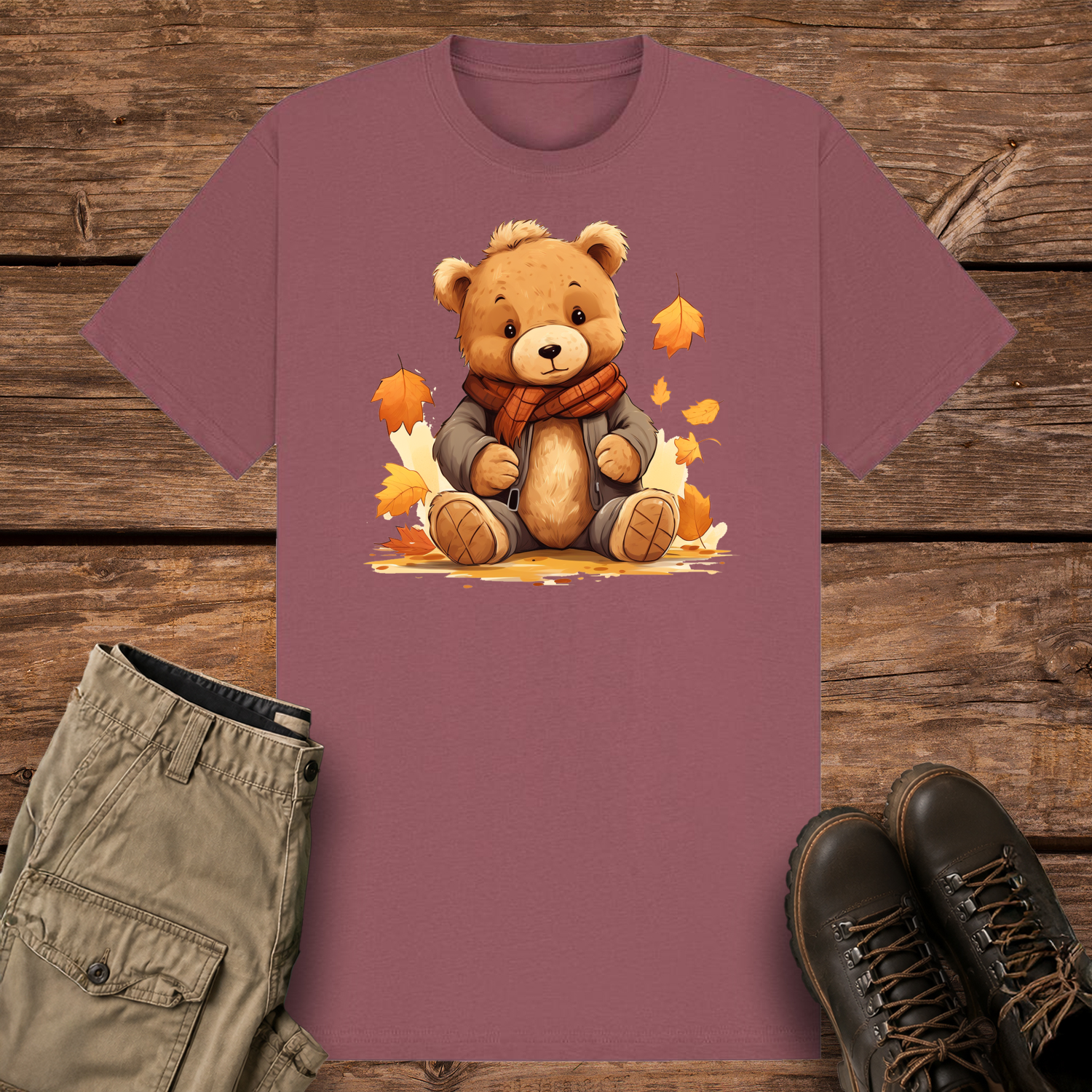 A mauve graphic t-shirt featuring a cute cartoon illustration of a fluffy brown teddy bear wearing a grey winter jacket and an orange plaid scarf. The bear is sitting surrounded by falling orange and yellow autumn leaves.