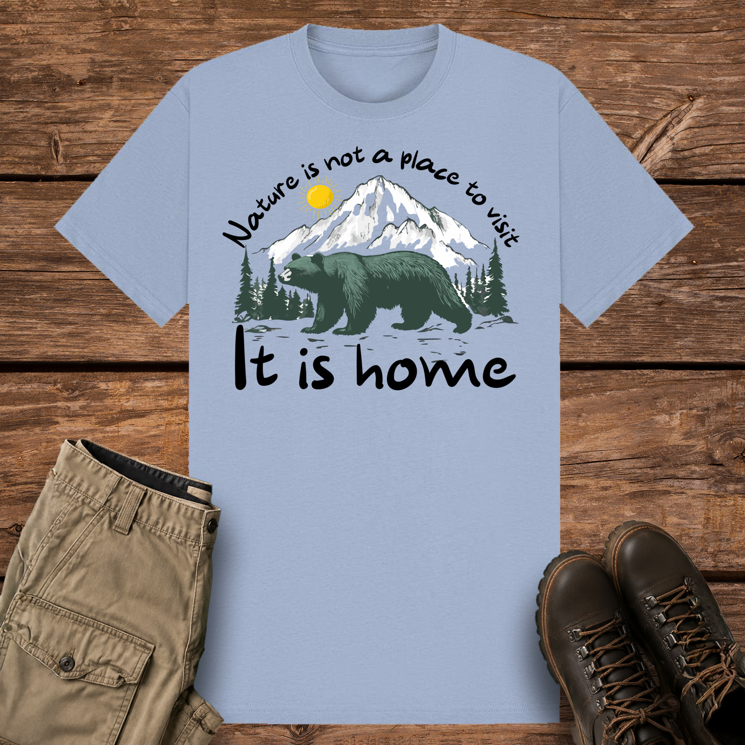 A pastel blue-colored graphic t-shirt featuring an illustration of a large dark green bear in front of snow-capped mountains and pine trees under a yellow sun. The text reads "Nature is not a place to visit" at the top and "It is home" at the bottom.
