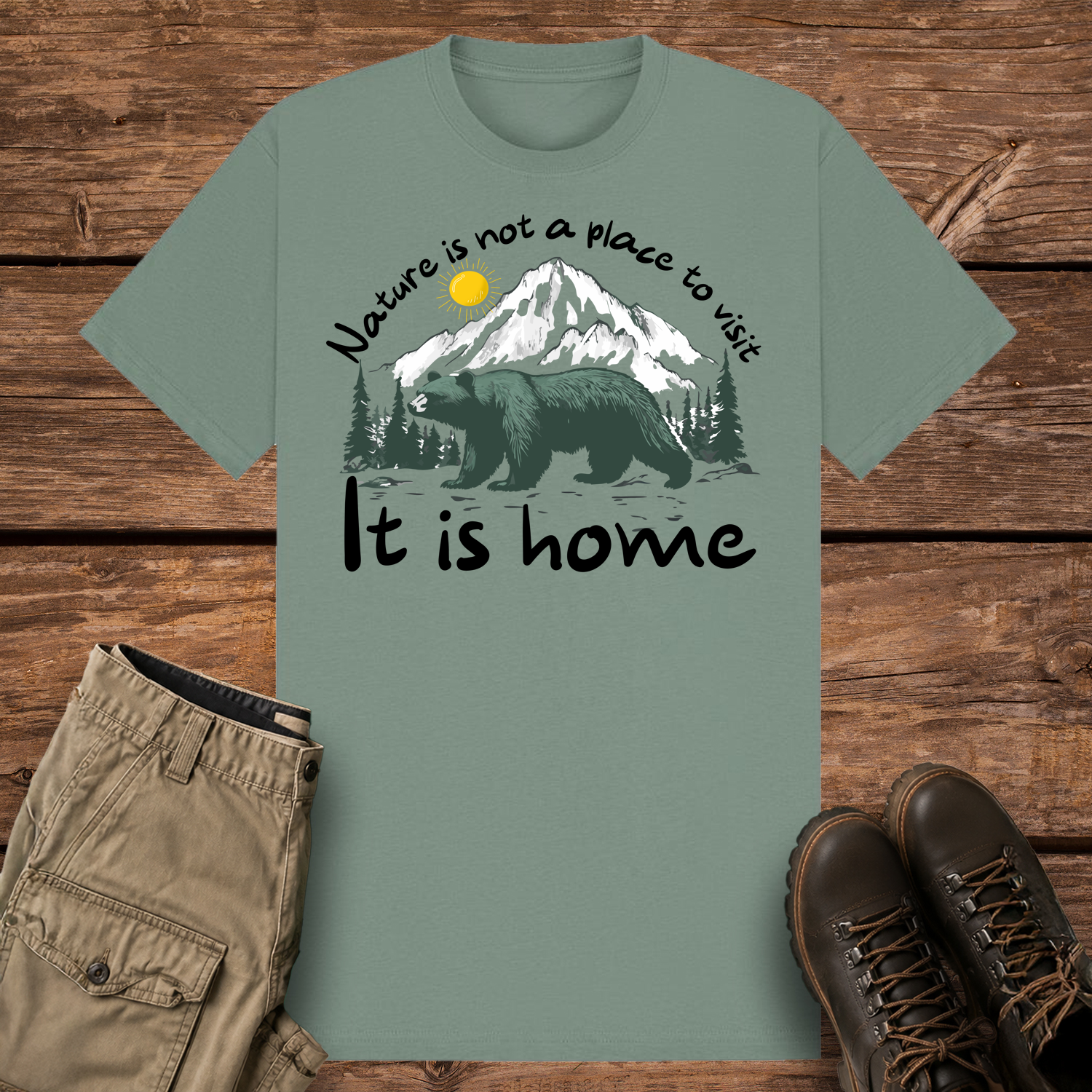 A sage-colored graphic t-shirt featuring an illustration of a large dark green bear in front of snow-capped mountains and pine trees under a yellow sun. The text reads "Nature is not a place to visit" at the top and "It is home" at the bottom.