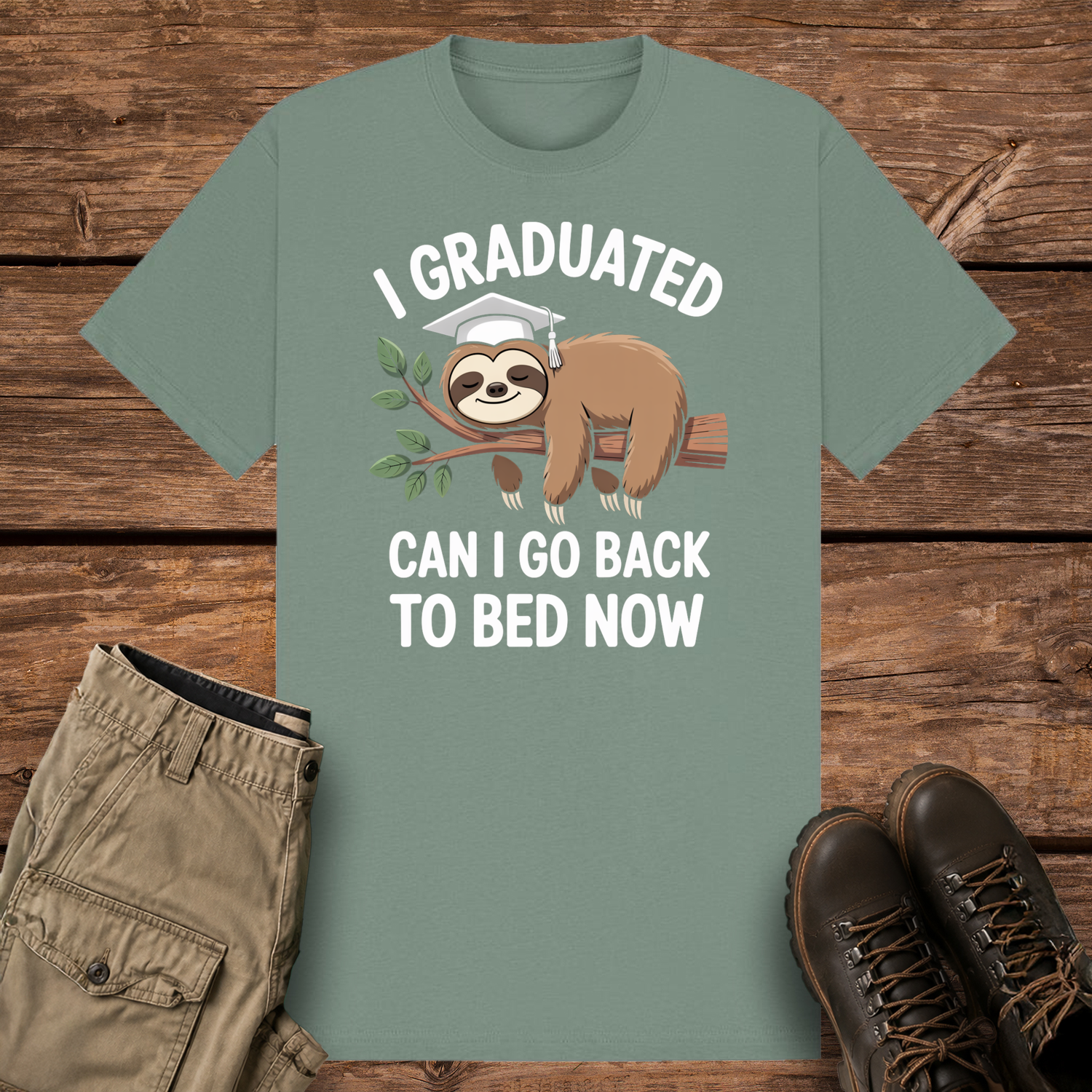 A sage graphic t-shirt featuring a cute cartoon illustration of a brown sloth wearing a white graduation cap while sleeping on a leafy tree branch. The white text reads "I GRADUATED" at the top and "CAN I GO BACK TO BED NOW" below.