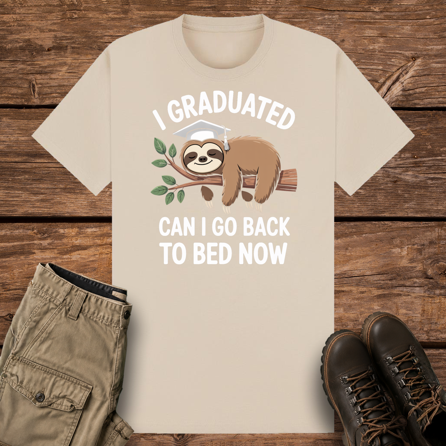 A sand graphic t-shirt featuring a cute cartoon illustration of a brown sloth wearing a white graduation cap while sleeping on a leafy tree branch. The white text reads "I GRADUATED" at the top and "CAN I GO BACK TO BED NOW" below.