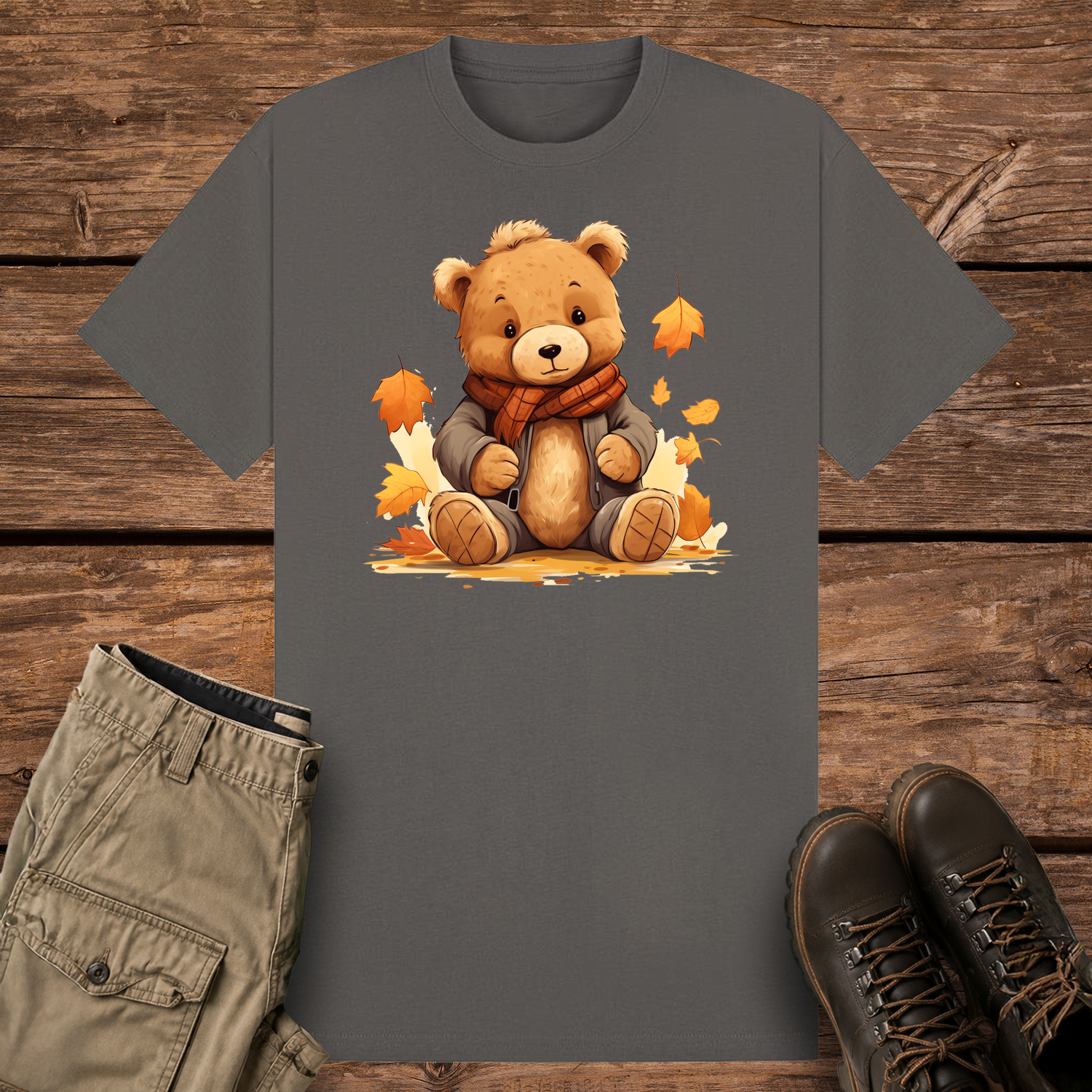 A charcoal graphic t-shirt featuring a cute cartoon illustration of a fluffy brown teddy bear wearing a grey winter jacket and an orange plaid scarf. The bear is sitting surrounded by falling orange and yellow autumn leaves.