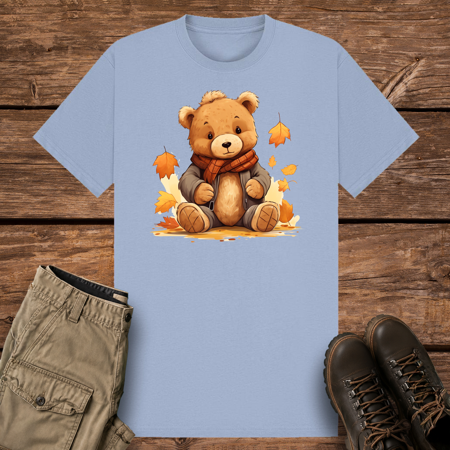A pastel blue graphic t-shirt featuring a cute cartoon illustration of a fluffy brown teddy bear wearing a grey winter jacket and an orange plaid scarf. The bear is sitting surrounded by falling orange and yellow autumn leaves.