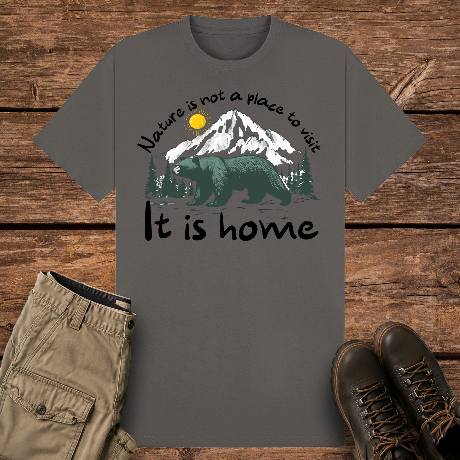 A charcoal-colored graphic t-shirt featuring an illustration of a large dark green bear in front of snow-capped mountains and pine trees under a yellow sun. The text reads "Nature is not a place to visit" at the top and "It is home" at the bottom.