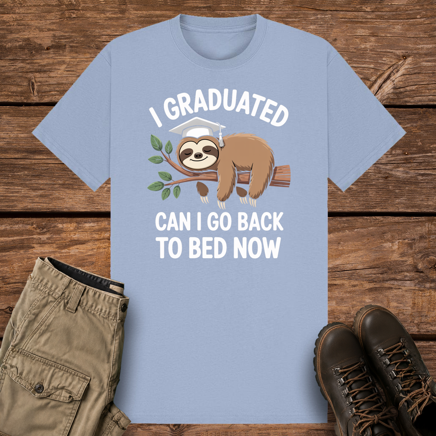 A pastel blue graphic t-shirt featuring a cute cartoon illustration of a brown sloth wearing a white graduation cap while sleeping on a leafy tree branch. The white text reads "I GRADUATED" at the top and "CAN I GO BACK TO BED NOW" below.