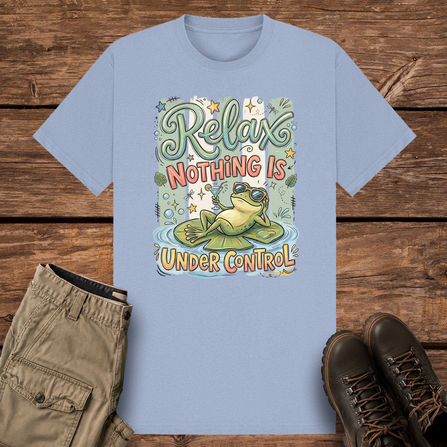 A pastel blue graphic t-shirt featuring a funny illustration of a relaxed green frog wearing sunglasses and holding a drink on a lily pad, surrounded by retro pastel stripes and the text "Relax Nothing Is Under Control''.
