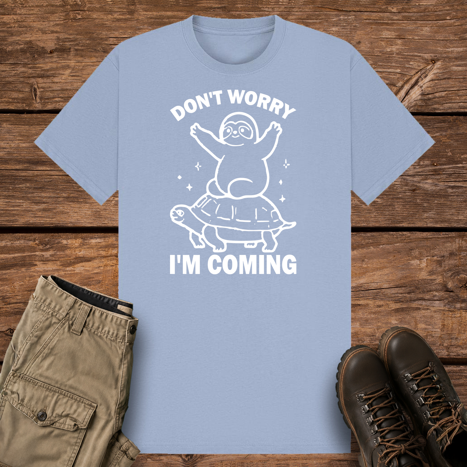 A pastel blue graphic t-shirt featuring a minimalist white line art design of a cute cartoon sloth sitting on top of a sea turtle's shell. The text reads "DON'T WORRY" at the top and "I'M COMING" at the bottom.