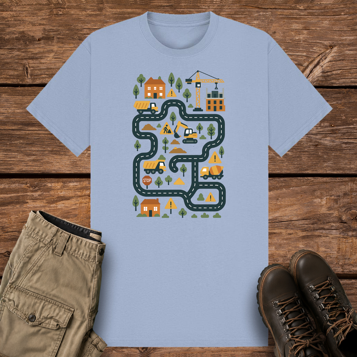 A pastel blue graphic t-shirt featuring a colorful illustration of a construction site road map. The design includes winding roads, cartoon dump trucks, an excavator, a crane, houses, trees, and yellow warning signs in a playful vector style.